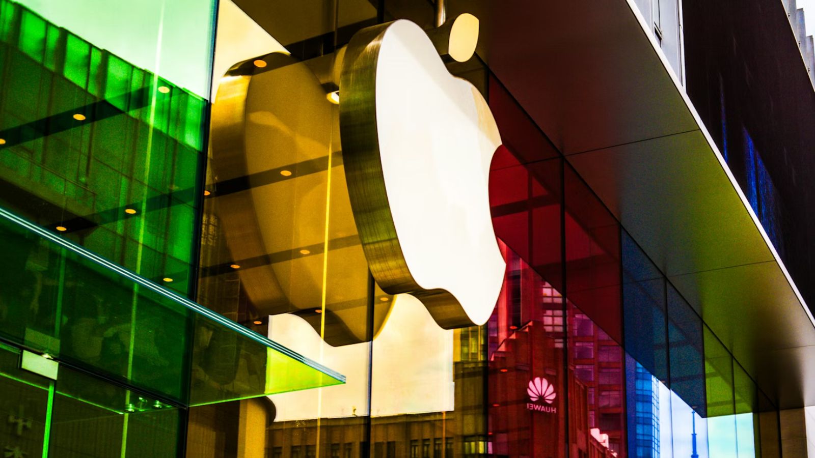 Fachada de uma loja da Apple com o icônico logotipo branco em destaque, refletido em painéis de vidro coloridos em tons de verde, amarelo e vermelho, em um ambiente urbano moderno.