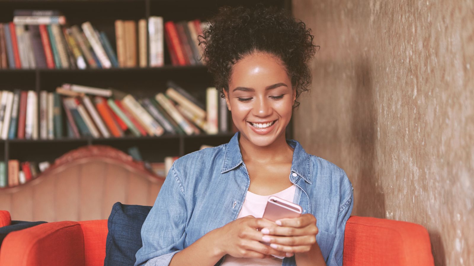 Mulher sorridente sentada em um sofá, usando o celular, com uma estante cheia de livros ao fundo, em um ambiente aconchegante que remete à leitura e ao acesso digital a conteúdos.