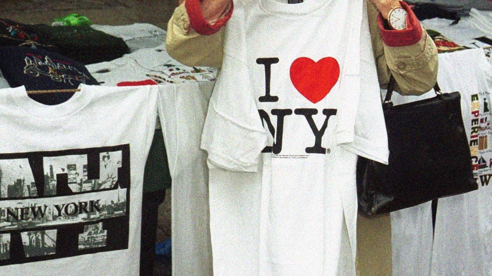 Pessoa segura uma camiseta branca com a frase “I ♥ NY” estampada em preto e vermelho. Outras camisetas brancas aparecem ao redor, sugerindo uma banca ou loja de souvenirs.