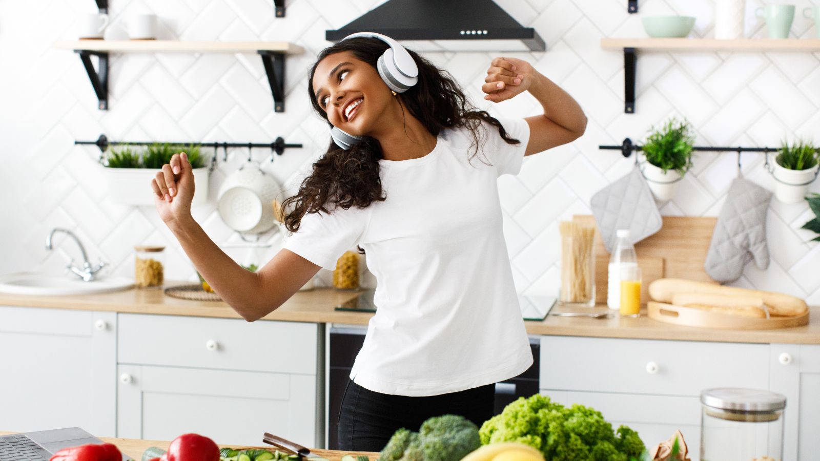 Mulher jovem em uma cozinha clara e organizada, usando fones de ouvido e dançando enquanto prepara alimentos.