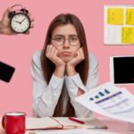 Mulher jovem sentada à mesa com expressão de cansaço e preocupação, apoiando o rosto nas mãos, enquanto várias mãos ao redor seguram objetos como relógio, celular, tablet, anotações e documentos, sugerindo excesso de tarefas e pressão por produtividade.