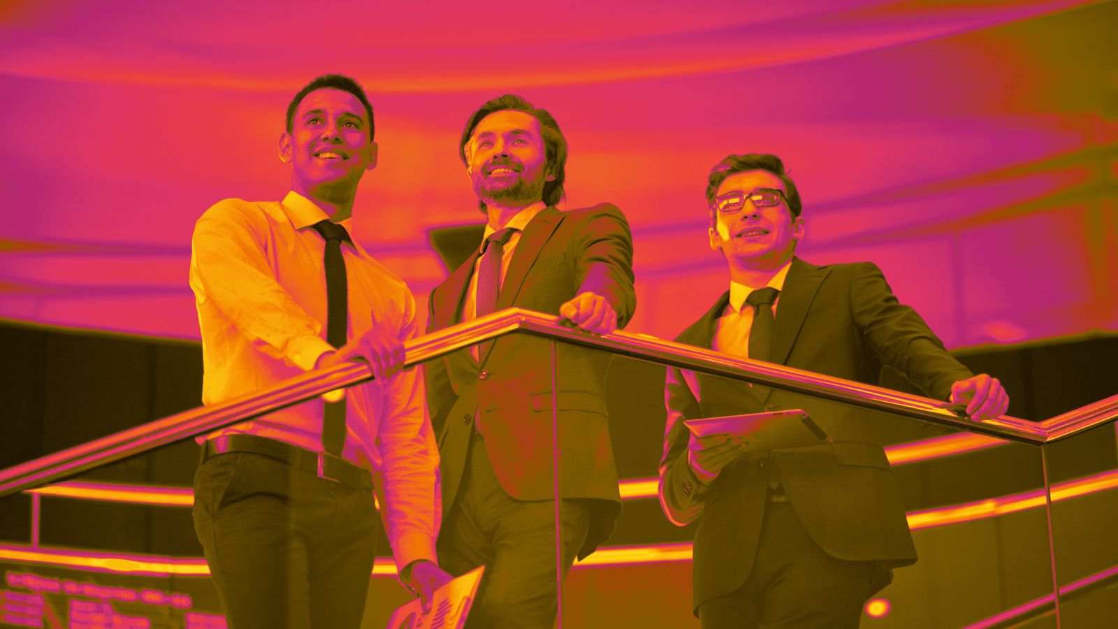 Três homens de terno estão apoiados em um corrimão dentro de um ambiente moderno, corporativo