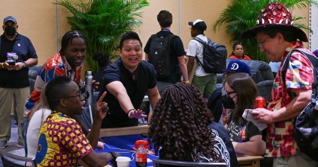 Participantes da Gen Con 2026 conversam e interagem ao redor de uma mesa durante o evento.