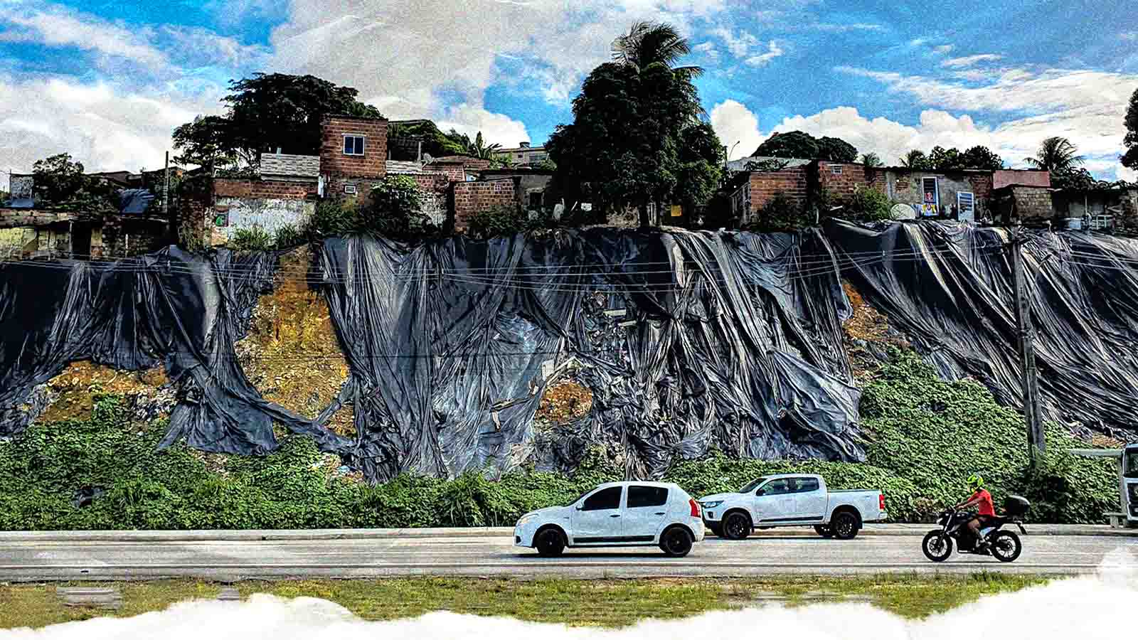 Casas de comunidade acima de uma encosta coberta por lonas pretas, ao lado de uma avenida
