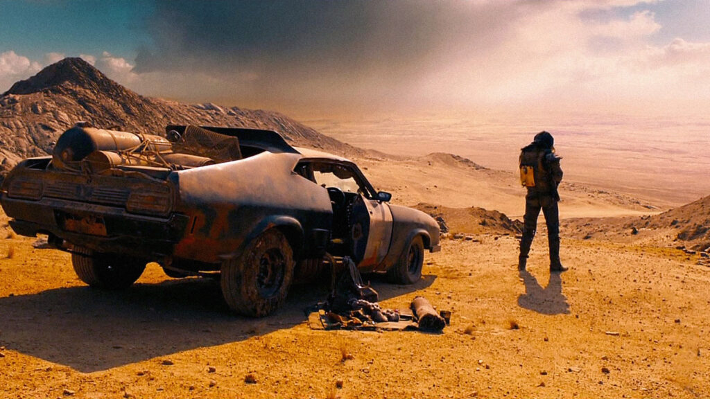 Homem observa uma paisagem des&eacute;rtica ao lado de um carro modificado na franquia Mad Max.