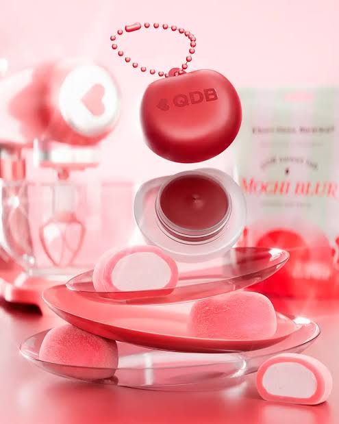 Embalagem vermelha do Mochi Blur aparece sobre potes de maquiagem e doces mochi em uma composi&ccedil;&atilde;o rosa.