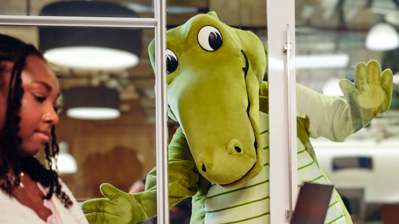 Niles, crocodilo verde e novo mascote da Crocs, aparece atrás de uma divisória de vidro em um escritório.