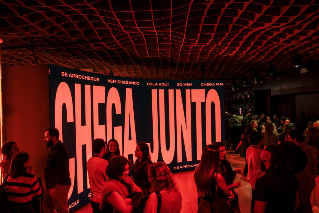Público circula pelo Behind the Streams diante de um painel vermelho com a frase “Chega junto”.