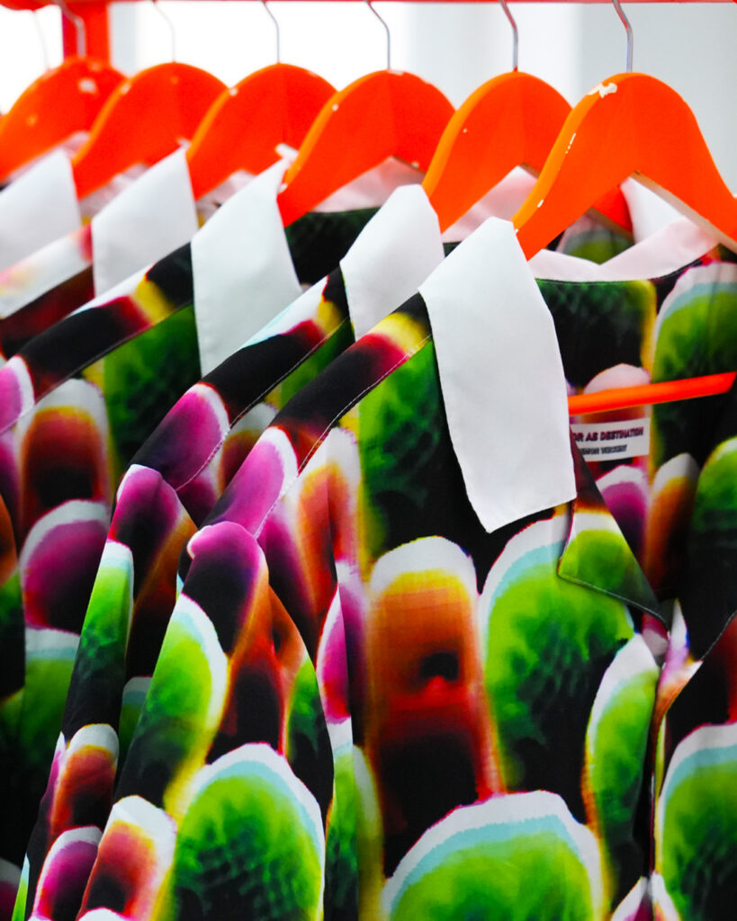 Camisas com estampas abstratas verdes, rosas e laranjas penduradas em cabides alaranjados.