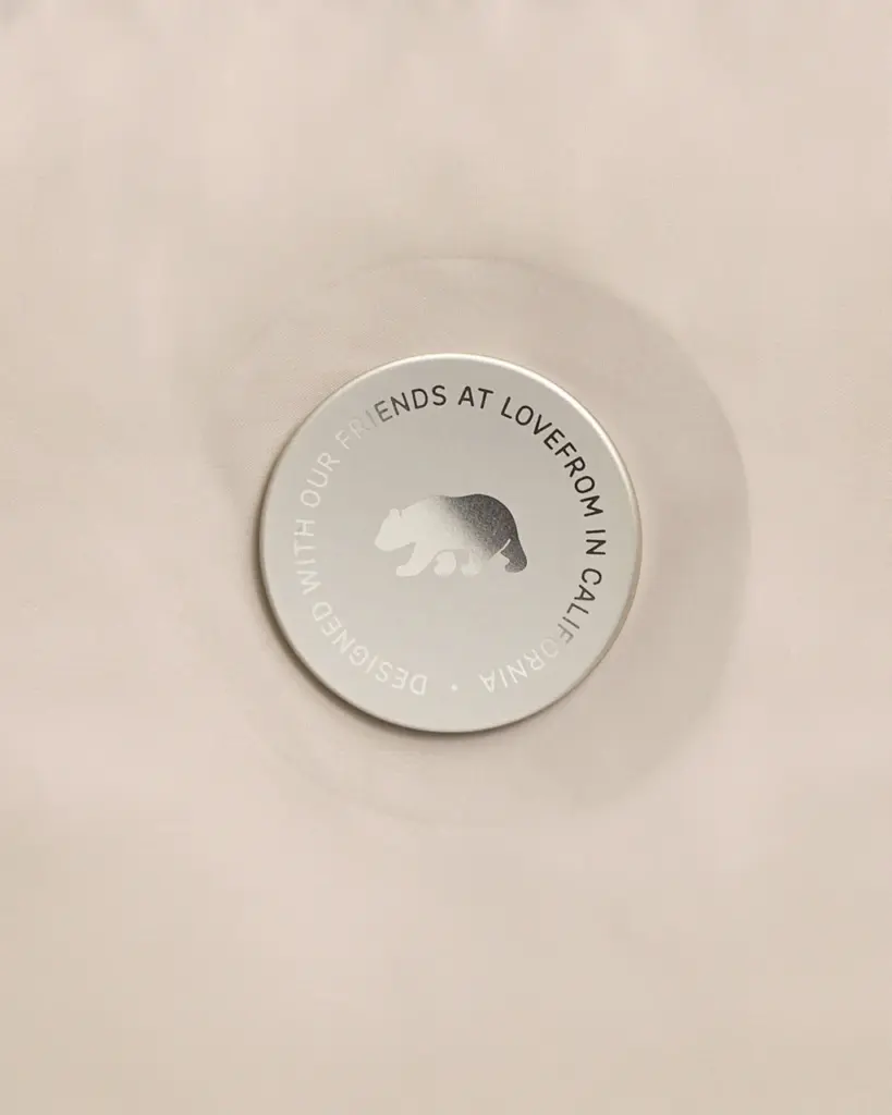Bot&atilde;o circular prateado sobre tecido claro, com o urso da LoveFrom ao centro e uma inscri&ccedil;&atilde;o ao redor.
