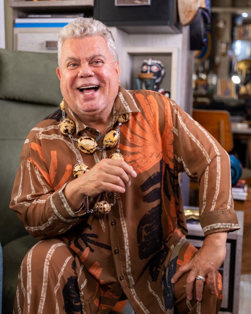 Milton Cunha sorri usando camisa de cetim estampada e um colar dourado, apoiado em uma divis&oacute;ria.
