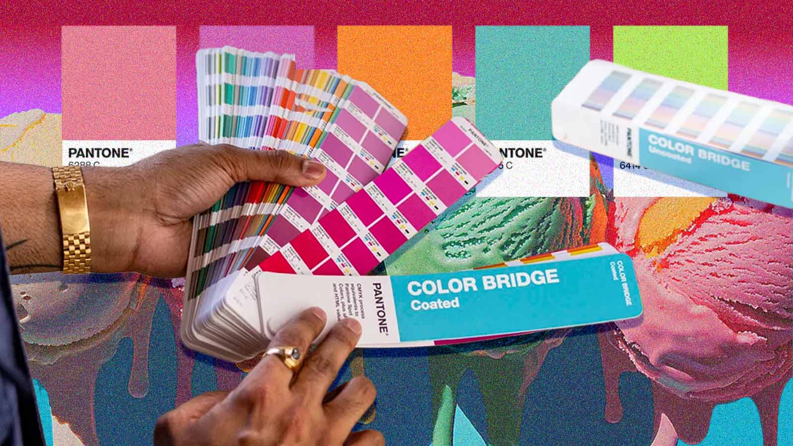 Mãos seguram leques de cores da Pantone sobre uma superfície com tons vibrantes.
