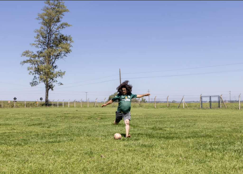 Pai de Danilo Zocatelli, maquiado e de peruca, chuta uma bola em um campo de futebol.