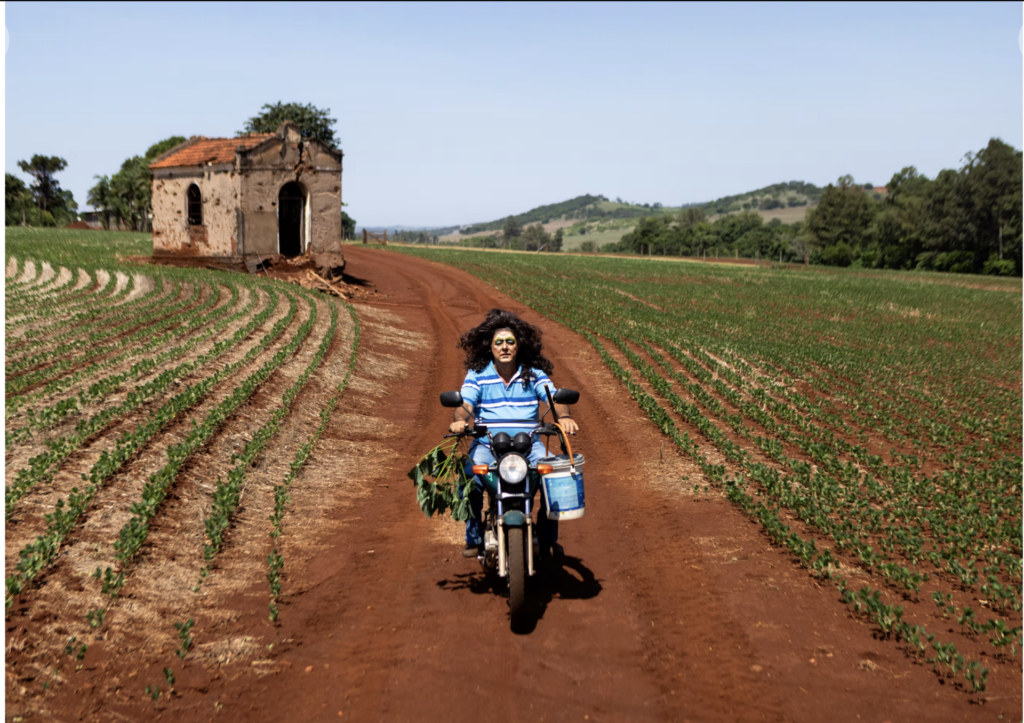 Pai de Danilo Zocatelli dirige uma motocicleta por uma estrada rural usando maquiagem e peruca.