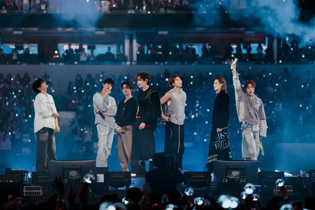 Integrantes do BTS reunidos no palco durante show da turn&ecirc; ARIRANG, diante de uma plateia com light sticks.