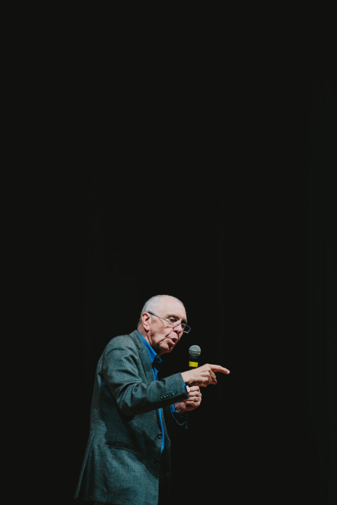 Gilles Lipovetsky segura um microfone e aponta com a mão durante uma palestra.