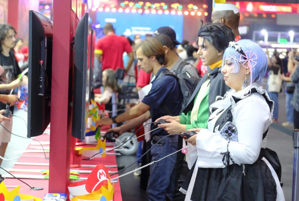 Dois cosplayers, um deles com peruca azul-clara, seguram controles diante de monitores de videogame, enquanto outros visitantes jogam ao lado.