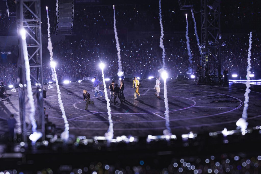 Integrantes do BTS se apresentam em palco cercado pelo público, com jatos de fumaça e luzes em tons de roxo.