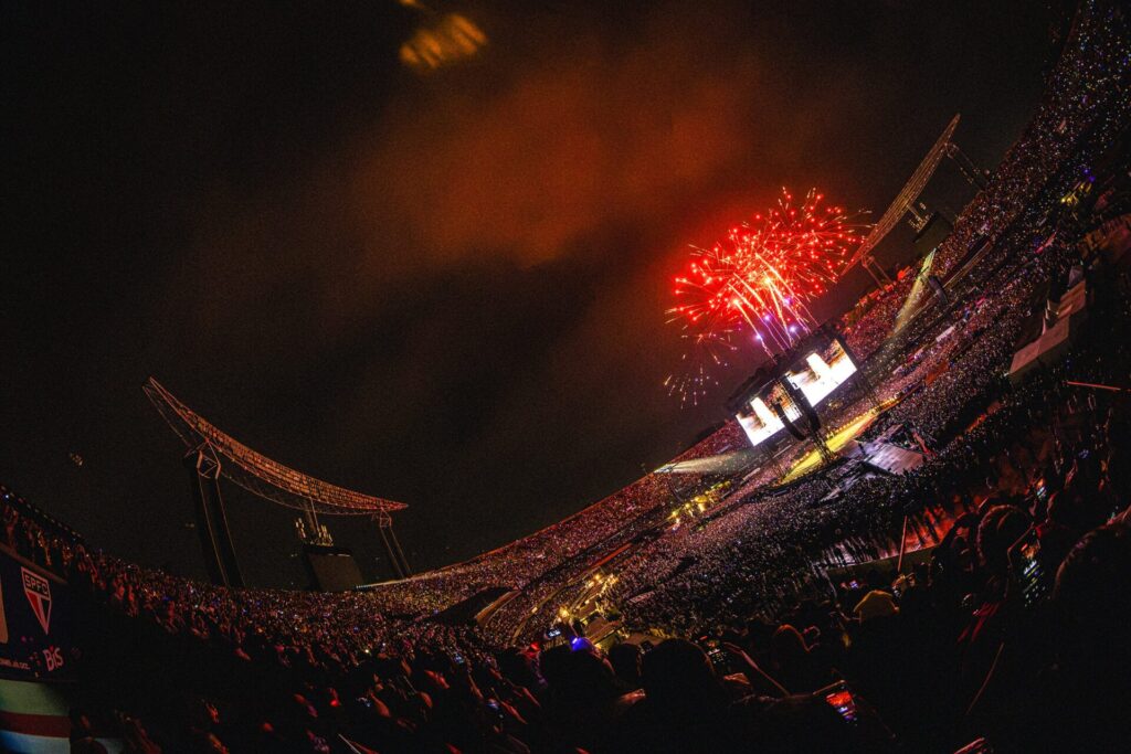 Vista panorâmica do MorumBIS lotado durante show do BTS, com fogos vermelhos sobre o palco.