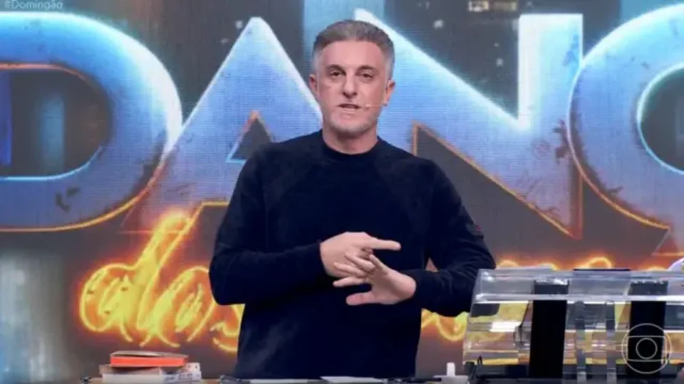 Luciano Huck, de camiseta preta e microfone no rosto, apresenta o Domingão diante do painel do programa.