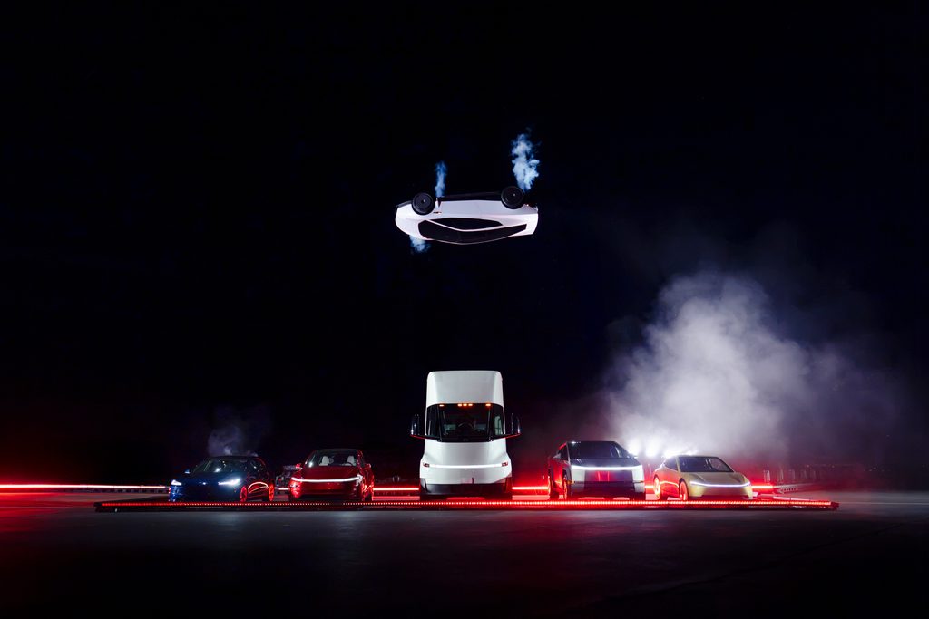 Tesla Roadster branco aparece de cabe&ccedil;a para baixo, suspenso no ar sobre uma fileira de ve&iacute;culos durante apresenta&ccedil;&atilde;o noturna.