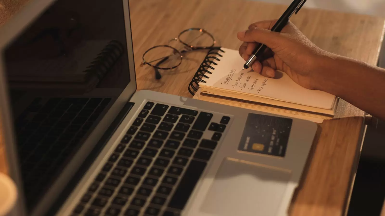 Mão escrevendo em um caderno ao lado de um laptop