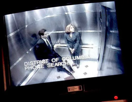 Uma captura de tela de um monitor de vigilância exibindo uma cena da série "Arquivo X". No interior de um elevador metálico, os agentes Mulder e Scully são vistos de um ângulo superior (plongée). Mulder está sentado no chão, encostado no canto, enquanto Scully está em pé ao lado dele, olhando para cima. Sobreposta à imagem, há uma legenda em letras brancas que diz: "DISTRICT OF COLUMBIA PHONE SEARCH". A moldura do monitor físico e uma pequena luz vermelha no canto inferior direito são visíveis.