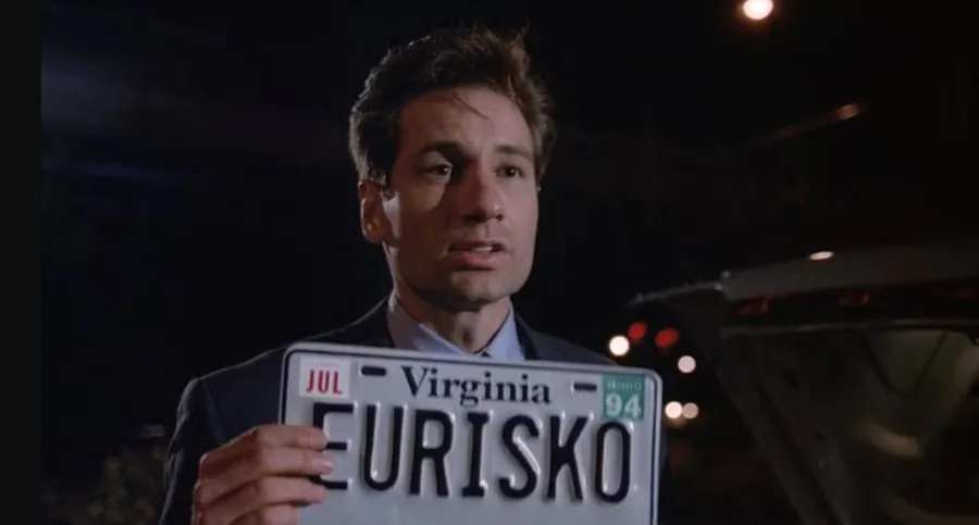 Um close do personagem Fox Mulder (interpretado por David Duchovny), da série "Arquivo X", em uma cena noturna. Ele segura em frente ao corpo uma placa de carro do estado da Virgínia, datada de julho de 1994, com a palavra "EURISKO" escrita em letras grandes e pretas. Mulder tem uma expressão de seriedade e surpresa.