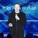 A imagem mostra Elon Musk em pé no centro de um palco, falando ao microfone durante uma apresentação. Ele veste um blazer preto texturizado sobre uma camiseta preta. Ao fundo, há um grande painel digital em tons de azul com design futurista, exibindo a palavra "TERAFAB" em letras grandes e brancas, acompanhada pelos logotipos da Tesla e da SpaceX em menor escala logo abaixo. Musk segura o microfone com a mão direita e gesticula levemente com a esquerda. Na parte inferior da foto, é possível ver a silhueta da plateia de costas, sugerindo o ambiente de uma conferência ou evento de tecnologia.