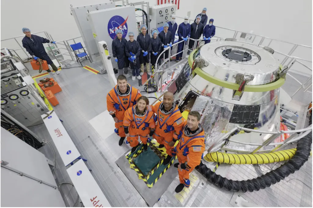astronautas da Artemis II, usando macac&atilde;o laranja, enfileirados.