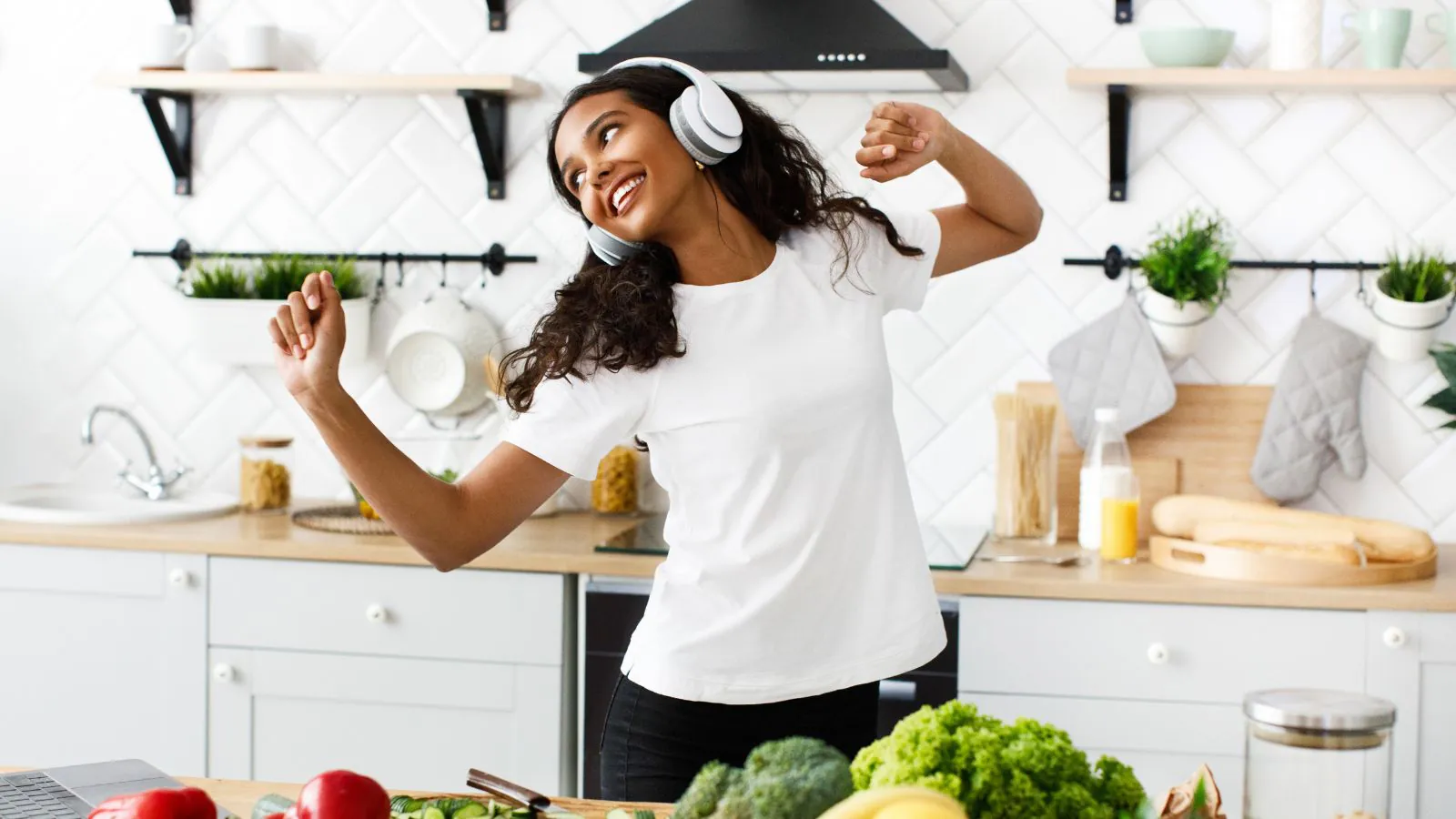 Mulher jovem em uma cozinha clara e organizada, usando fones de ouvido e dançando enquanto prepara alimentos.