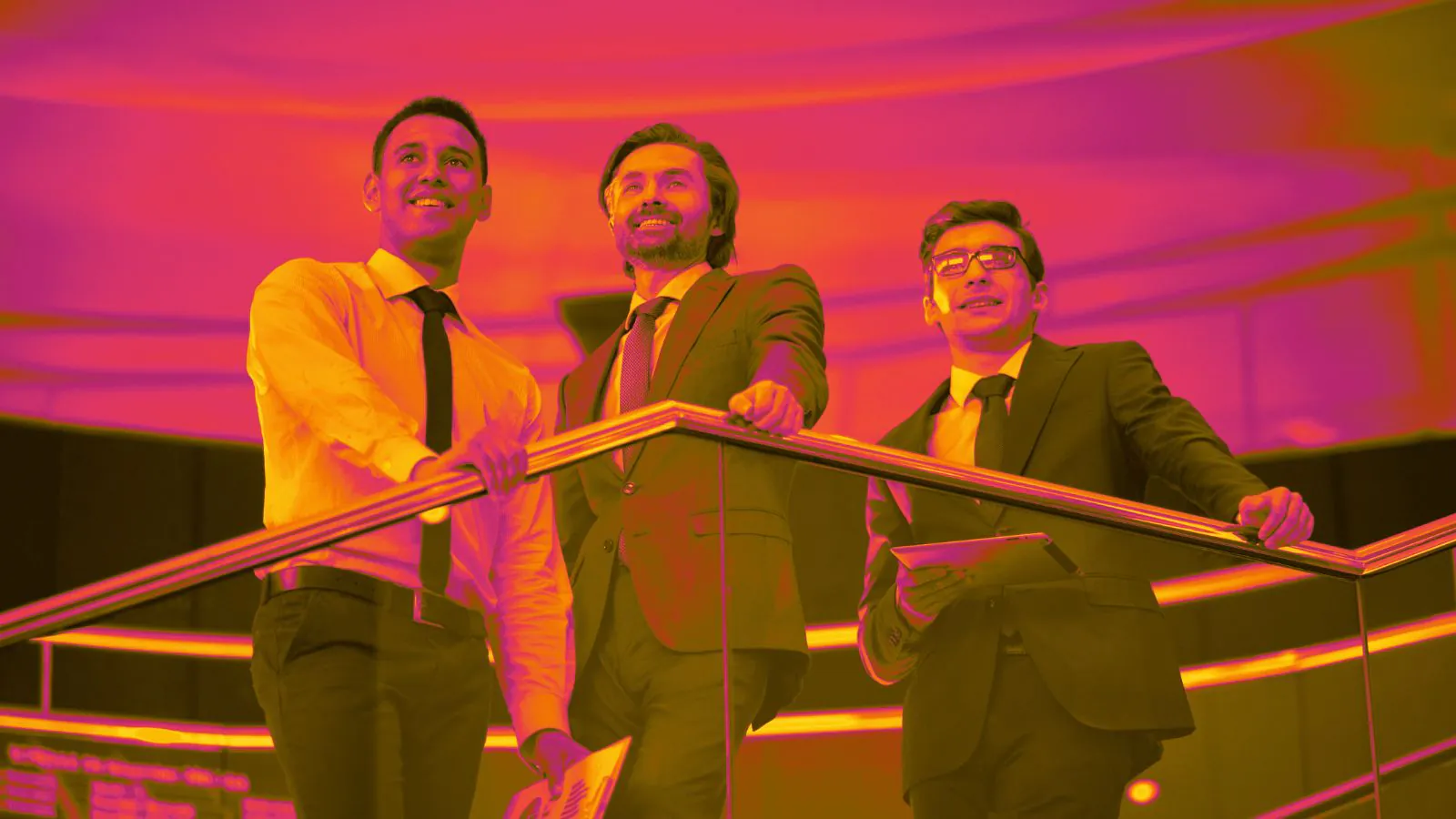 Três homens de terno estão apoiados em um corrimão dentro de um ambiente moderno, corporativo