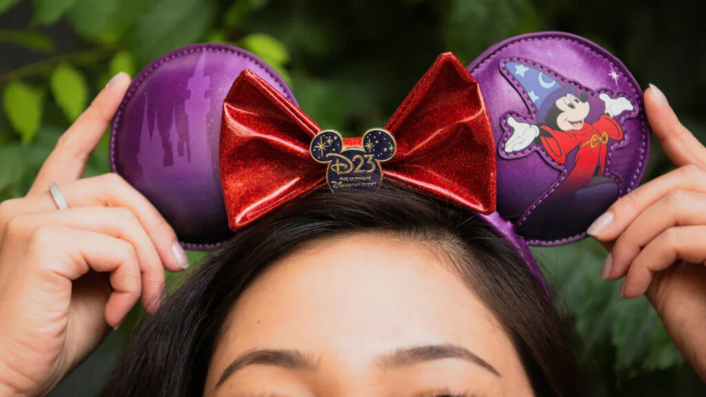 Pessoa usa tiara roxa com orelhas de Mickey, laço vermelho e símbolo do D23 2026.