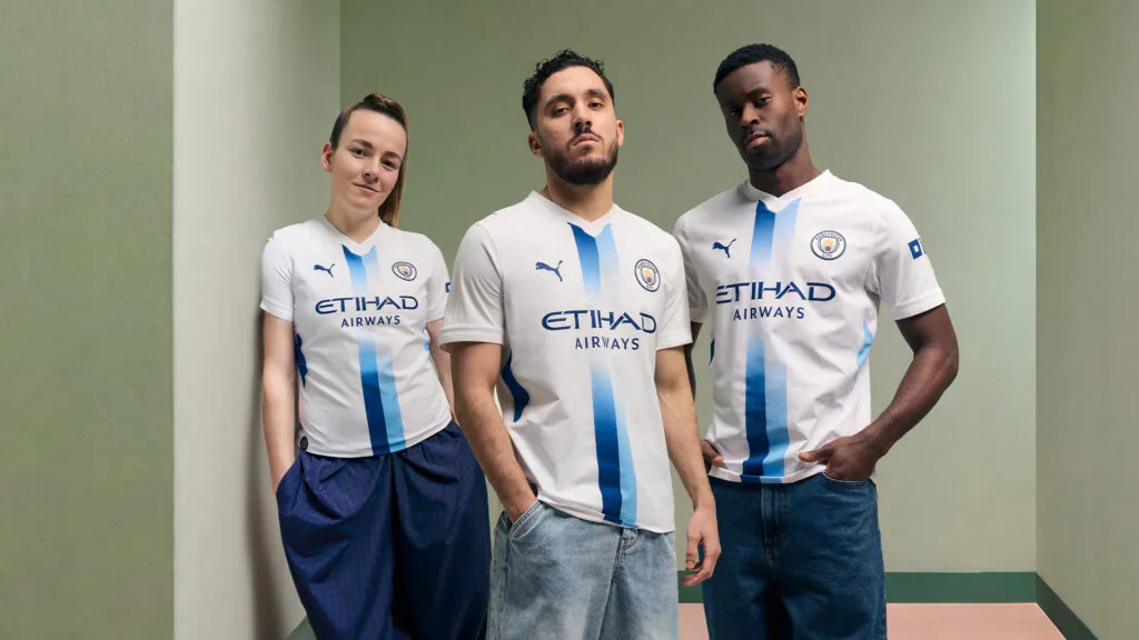 Tr&ecirc;s atletas do Manchester City posam com o uniforme branco criado por torcedores com intelig&ecirc;ncia artificial.