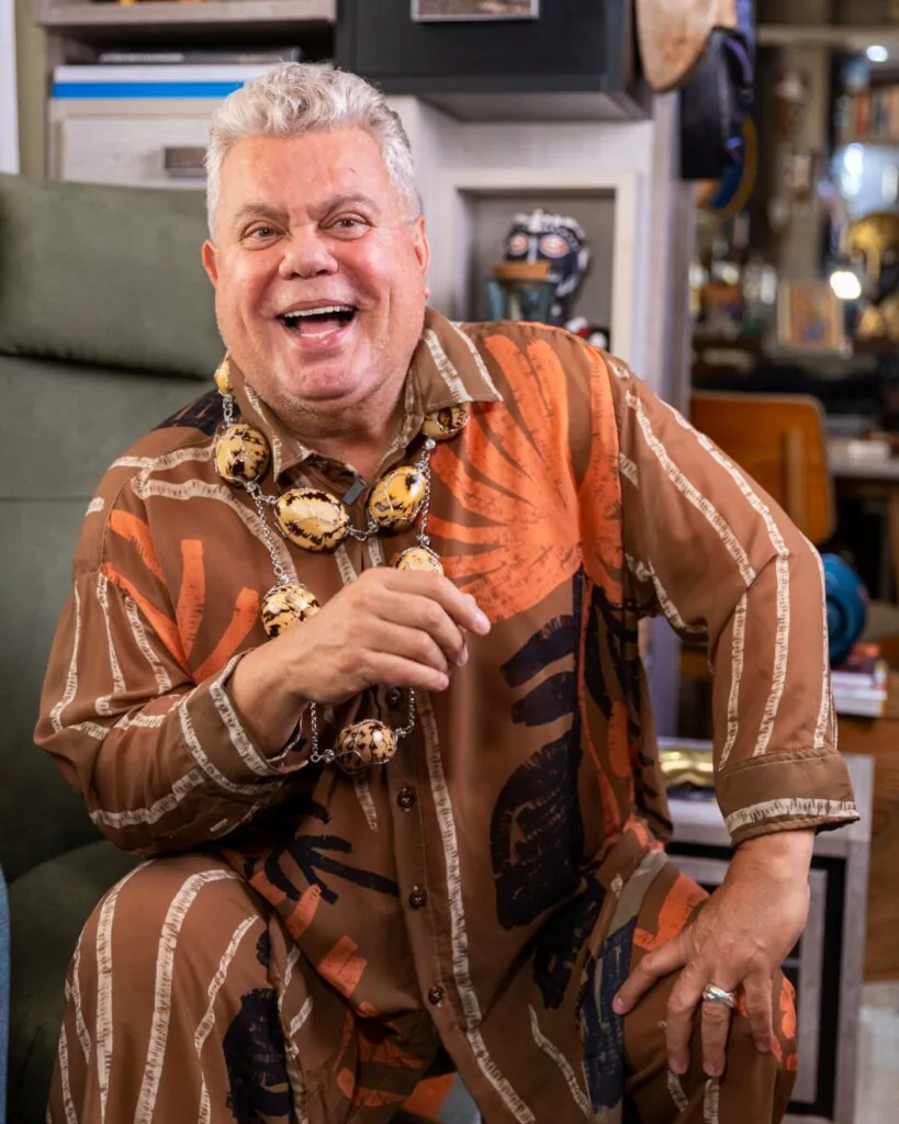 Milton Cunha sorri usando camisa de cetim estampada e um colar dourado, apoiado em uma divisória.