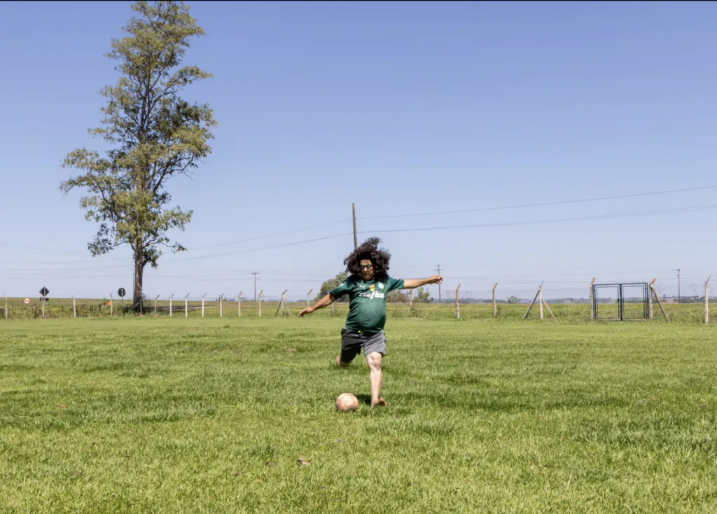 Pai de Danilo Zocatelli, maquiado e de peruca, chuta uma bola em um campo de futebol.