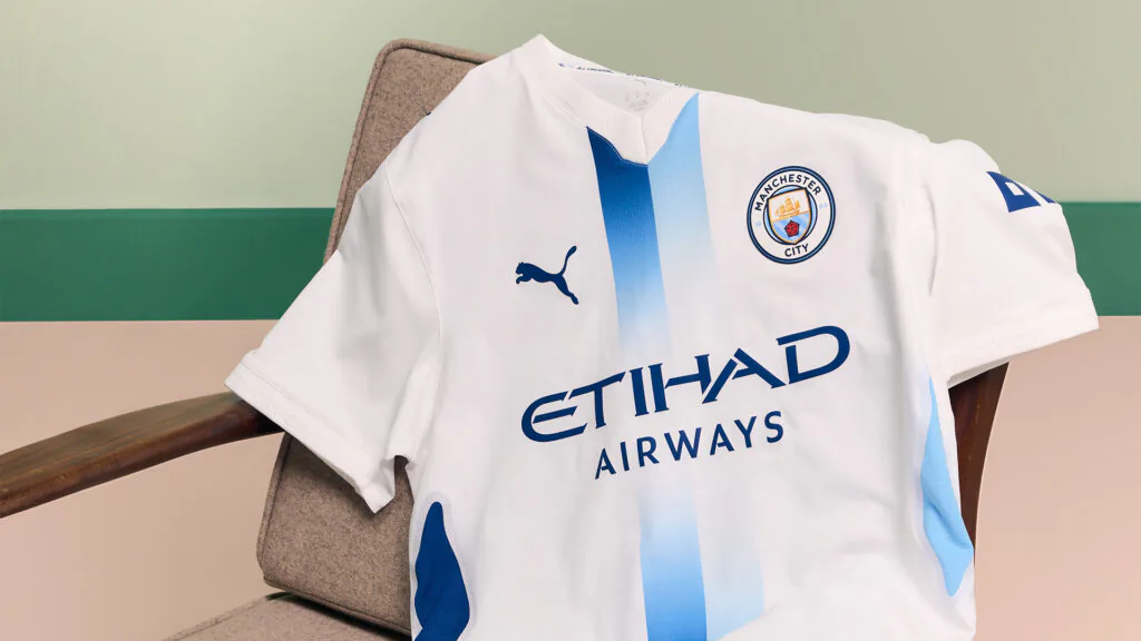 Camisa branca do Manchester City com duas faixas verticais em azul-claro e azul-marinho, escudo do clube e logotipo da Puma.