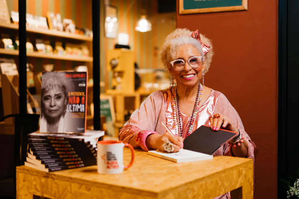 S&ocirc;nia Guimar&atilde;es sorri sentada &agrave; mesa em uma livraria, com exemplares de seu livro "A primeira, n&atilde;o a &uacute;ltima" ao fundo.